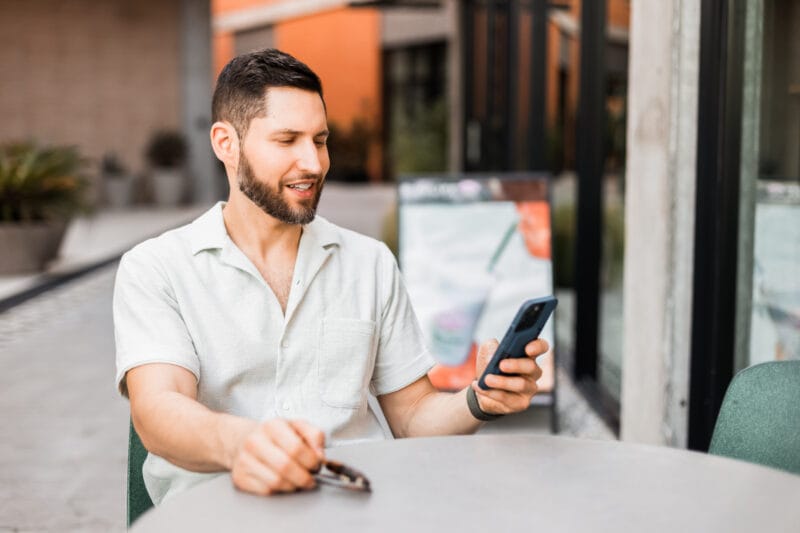 David Shapiro seated at an outdoor cafe table, smiling while looking at his phone